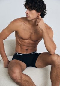 Crne bokserice s bijelim pojasom na kojem piše "BORN BORN." Model ima vitku tjelesnu građu i sjedi na svijetlo obojenoj teksturiranoj stolici.