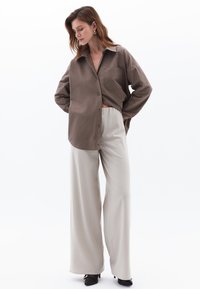 Camicia oversize marrone con bottoni, dotata di tasca, abbinata a pantaloni crema a gamba larga. L'outfit presenta un tessuto morbido e una vestibilità comoda.