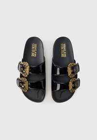 Crne lakirane sandale na sklizanje s dva remena sa kopčama ukrašenim raskošnim zlatnim detaljima. Teksturirana potplata s logotipom "Versace Jeans Couture".