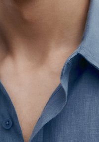 Ljusblå skjorta med krage i en texturerad tyg, synliga knappar och en prydligt vikt krage. Närbild av halsområdet.