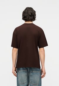 Άτομο με κοντά σγουρά σκουρόχρωμα μαλλιά που φοράει απλό σκούρο καφέ κοντομάνικο μπλουζάκι και ανοιχτόχρωμο μπλε τζιν, με την πλάτη προς την κάμερα.