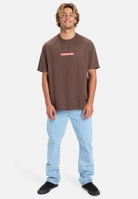 Brun bomulds T-shirt med et rødt og hvidt "Quiksilver" logo, parret med lyseblå jeans og sorte slip-on sko. Lige skåret design.