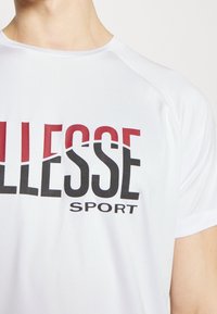 Camiseta deportiva blanca con un texto audaz y contrastante en negro y rojo que dice "ELLESSE SPORT." Está hecha de una tela suave y ligera con cuello redondo.