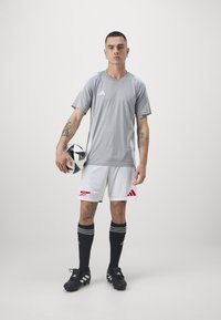 Camiseta atlética gris con acentos blancos, pantalones cortos blancos con logo rojo, calcetas negras con rayas blancas y botas de fútbol negras, sosteniendo un balón de fútbol.