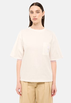 Donna con capelli scuri lisci indossa una t-shirt oversize color crema con tasca e pantaloni beige a gamba larga su sfondo grigio chiaro.