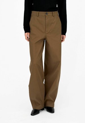 Personne portant un pantalon marron taille haute à jambes larges avec poches avant et des chaussures noires pointues, debout sur fond blanc.