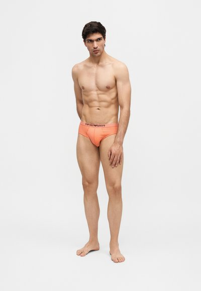 Νέος άνδρας με γυμνασμένο σώμα, ντυμένος με φωτεινά πορτοκαλί εσώρουχα Emporio Armani, όρθιος ξυπόλητος σε απλό ανοιχτόχρωμο φόντο.