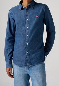 Donkerblauw denim overhemd met een button-down kraag, lange mouwen en witte knopen. Bevat een rood Levi's logo op de borstzak.