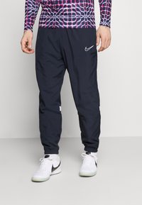 Man som bär marinblå Nike träningsbyxor med elastiska handledsfästen, vita Nike sneakers och en mönstrad långärmad skjorta, stående framför en enhetlig bakgrund.