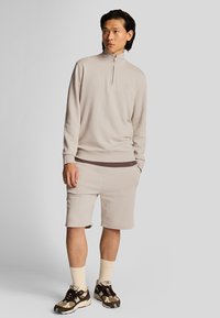 Beige zip-up sweatshirt met een staande kraag, gecombineerd met beige shorts. Beide gemaakt van een gladde stof, met een subtiele logo en geribbelde accenten.