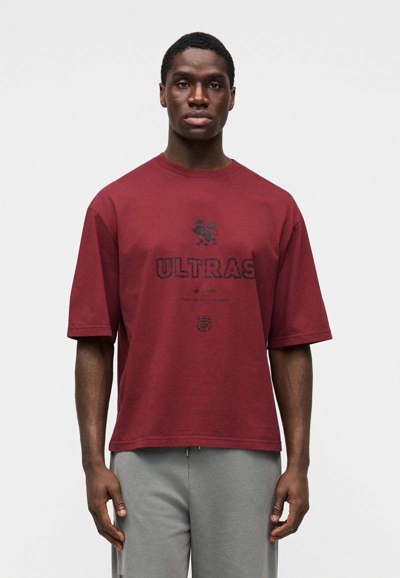 T-shirt de algodão cor castanha avermelhada com mangas curtas, apresenta o texto "ULTRAS" e um gráfico de leão a preto. Bainha reta e corte relaxado.
