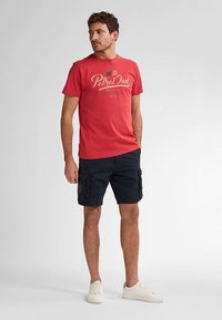 Red cotton t-shirt with graphic print and crew neck, paired with navy cargo shorts featuring pockets, worn with white sneakers.