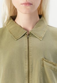 Olijfgroene ritsjack met grote kraag, voorzak en gestructureerde stof, met zichtbare stiksels en een gestroomlijnd ontwerp.