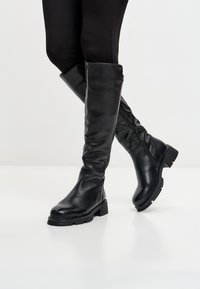 Gambe che indossano leggings neri e stivaletti neri di pelle alti fino al ginocchio con tacco basso e chunky, in piedi su un pavimento bianco contro uno sfondo neutro.