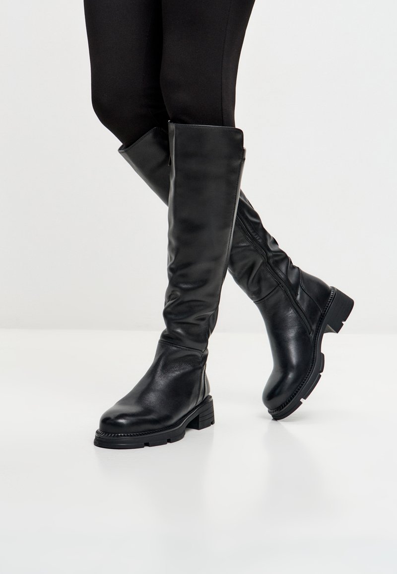 Gambe che indossano leggings neri e stivaletti neri di pelle alti fino al ginocchio con tacco basso e chunky, in piedi su un pavimento bianco contro uno sfondo neutro.