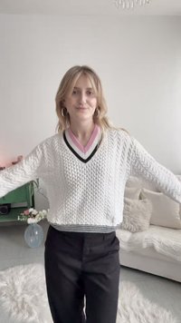 Νεαρή γυναίκα με ξανθά μαλλιά που φοράει λευκό πουλόβερ πλεκτό με σχέδιο και μαύρο παντελόνι, στέκεται σε ένα φωτεινό σαλόνι με ένα βάζο με λουλούδια.