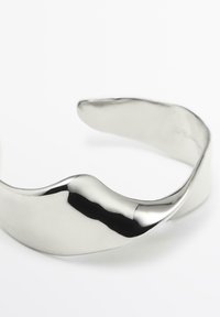 Zilveren manchetarmband met een glad, golvend ontwerp. Gepolijste metalen afwerking weerspiegelt het licht en benadrukt de krommingen en contouren. Geen zichtbare sluitingen.