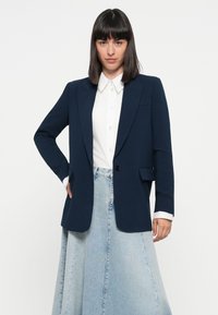 Marineblazer med én knapp, notchklaffer og frontlommer, kombinert med en hvit skjorte og et lyst denimskjørt. Glatt stofftekstur.