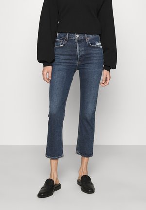 Model wearing dark blue cropped flare jeans with slight distressing near pockets, paired with black long-sleeve top and black slip-on shoes.