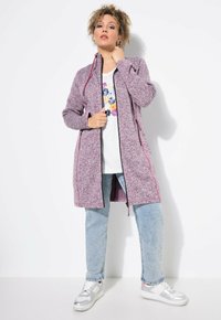Lilla, struktureret lang cardigan med sort lynlås og røde detaljer, båret over en hvid blomstret t-shirt og lyseblå jeans. Sølvfarvede sneakers.