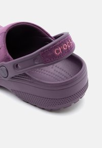 Sabots Crocs violets avec sangle réglable, ornés d'un logo brodé et de semelles texturées, vus d'un angle rapproché arrière.