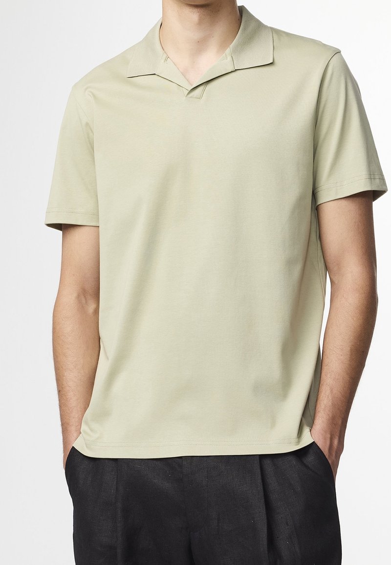 Homme portant un polo beige à manches courtes avec col et un pantalon noir, les mains dans les poches, devant un fond uni.