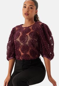 Burgundifärgad texturerad topp med puffärmar, som har 3D-blomdekorationer och ett semitransparent tyg, i kombination med åtsittande svarta byxor.