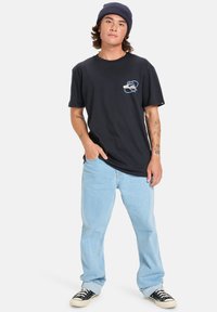 Tricou negru din bumbac cu un grafic albastru și bej pe piept, purtat cu blugi deschiși la culoare și adidași negri. Potrivire casual și stil relaxat.
