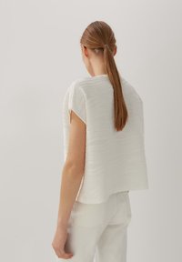 Kvinde med lang brun hestehale iført en struktureret hvid kortærmet top og hvide bukser, vist bagfra mod en neutral baggrund.