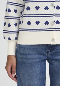 Cárdigan blanco con estampados de corazones azules, rayas horizontales, puños de canalé y cinco botones, combinado con vaqueros azules de mezclilla.