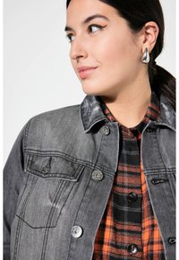 Harmaa farkkutakki, jossa on musta kaulus, hopeiset yksityiskohdat ja rintapocket. Päällä punavalkoraidallinen flanellipaita.