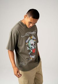Olive green short-sleeve t-shirt with a skull and heart graphic, "Ed Hardy" logo, and colorful stitching details. Cotton fabric, relaxed fit.