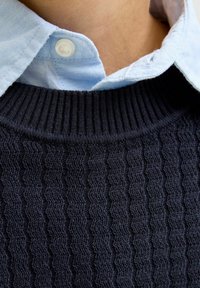 Detailní záběr na svetr s námořnickou modrou kabelkovou vazbouovou texturou, který je umístěn na límci světle modré košile na krku osoby.