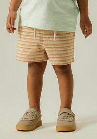 Shorts rayés en crème avec des rayures orange, dotés d'une taille avec cordon de serrage. Associés à des chaussures en suède beige avec des semelles en caoutchouc.