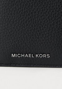 Fekete textúrájú bőr felület ezüstszínű "MICHAEL KORS" márkanevével, amely a alsó szélén van bemélyítve.