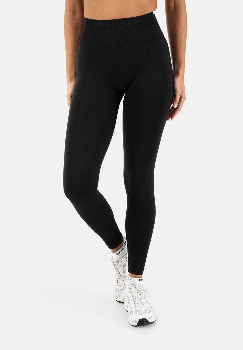 Femme portant des leggings noirs taille haute et des chaussures de sport blanches, debout avec une jambe croisée sur l'autre sur un fond uni.