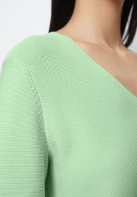 Green knitted sweater with a deep V-neck and textured fabric, featuring ribbed detailing along the edges. Close-up view of the neckline.