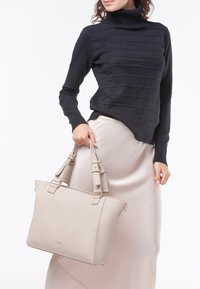 Borsa in pelle grigia con doppi manici e hardware dorato. Presenta una superficie testurizzata e una forma rettangolare, tenuta da una persona che indossa un maglione scuro.