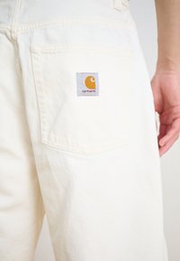 Beli denim hlače z zadnjim žepom in kvadratno, oranžno nalepko Carhartt. Gladka tekstura, dizajn naravnost nogavic.