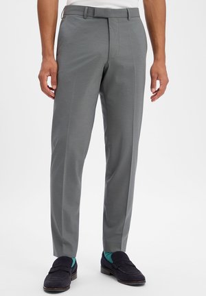 Man met grijze op maat gemaakte broek, donkerblauwe suède loafers en teal met patroon sokken, staand tegen een witte achtergrond.