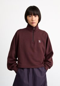 Sudadera de media cremallera color burdeos con un cuello alto, hombros caídos y un logo bordado en el pecho, combinada con pantalones plisados.