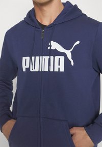 Námořnická mikina na zip vyrobená z bavlněné směsi. Uprostřed hrudi se nachází velké bílé logo "PUMA" a silueta skákající pumy. Má přední kapsy.