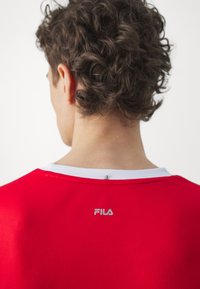 Camiseta atlética roja con una textura de malla, cuello blanco y un pequeño logotipo plateado de Fila en la parte posterior. Cabello rizado visible en la parte superior.