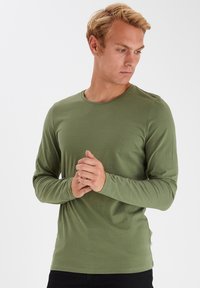 Ung mand med kort blondt hår, iført en tætsiddende olivengrøn langærmet skjorte, som klapper hænderne og ser ned til siden.