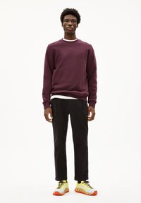 Felpa girocollo bordeaux con dettagli bianchi al collo, pantaloni neri e sneakers colorate con accenti gialli, grigi e arancioni.