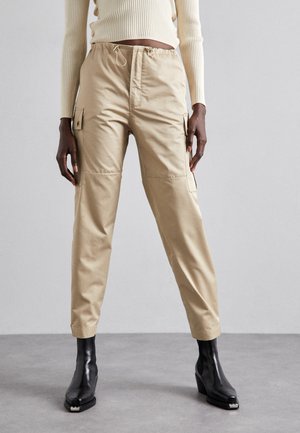 Beige cargobroeken met elastische tailleband en trekkoord, met zijzakken. Draagt men met zwarte hoge hakken, gladde textuur.