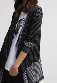 Svart och grå randig cardigan med öppen framsida, långa ärmar och en gradientdesign. Bärs över en vit t-shirt med tryck.