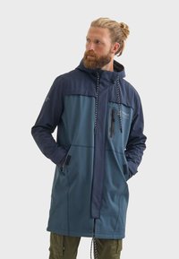 Homme avec une barbe portant une veste imperméable bicolore, dotée d'une capuche et de poches zippées, se tient devant un fond uni.