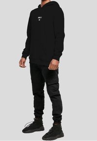 Felpa nera con la scritta "ASSOLUTAMENTE NO", tasca frontale, indossata con pantaloni cargo neri e scarpe sportive nere su uno sfondo grigio.