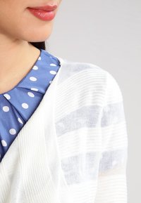 Vit stickad cardigan med randig textur, lagd över en blå blus med prickar. Blusen har en plisserad krage.